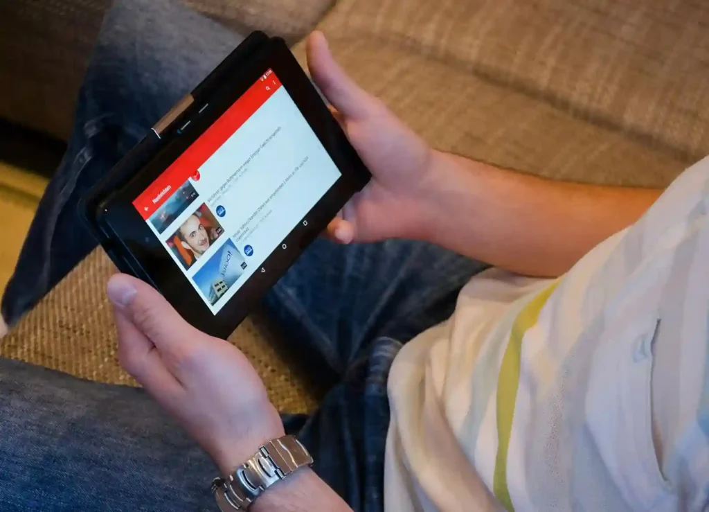 A person holding a tablet is viewing some thumbnails on a YouTube while sitting on a couch. The person is wearing a striped shirt and jeans.